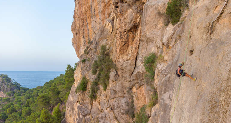Descripción de la foto de portada: En primer plano aparece una mujer rapelando la pared de una montaña con cuerda verde, casco y mochila. Al fondo se ve un mar azul y entre la roca y el mar un manto verde de árboles.