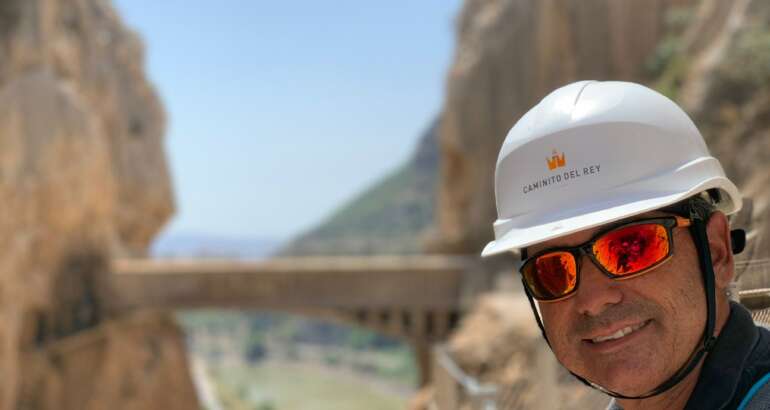Se ve en primer plano a Juan José López Rosa, nuevo responsable del Área de senderismo y senderos de la FEDME, con casco, sonriente, al fondo, desenfocado se una pasarela entre dos montañas.