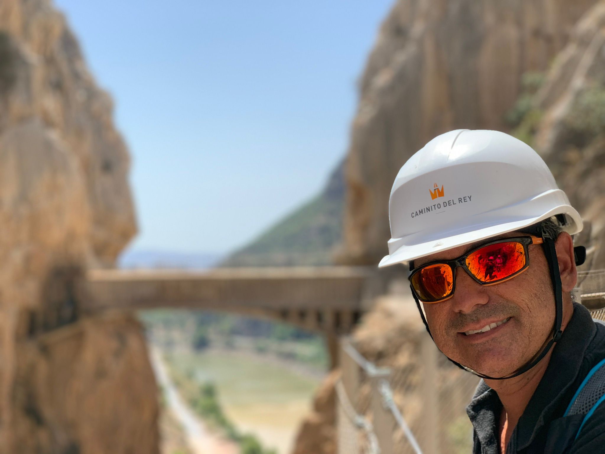 Se ve en primer plano a Juan José López Rosa, nuevo responsable del Área de senderismo y senderos de la FEDME, con casco, sonriente, al fondo, desenfocado se una pasarela entre dos montañas.