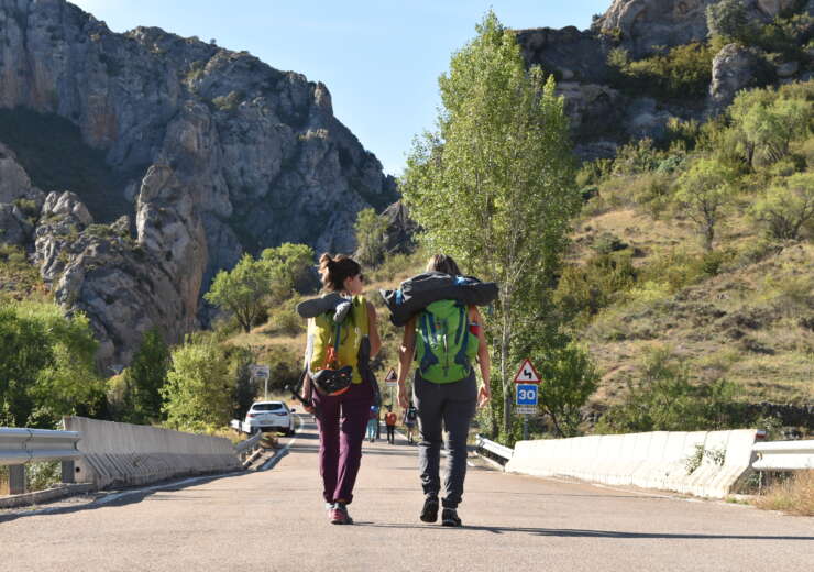 Aparecen dos chicas de espalda caminando por carretera hacia las montañas que se ven al fondo. Ambas llevan mochilas cargadas con útiles de escalada