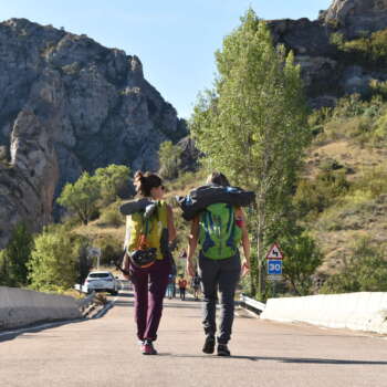 Aparecen dos chicas de espalda caminando por carretera hacia las montañas que se ven al fondo. Ambas llevan mochilas cargadas con útiles de escalada
