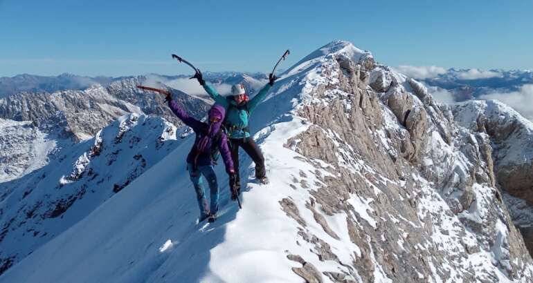 Dos mujeres alpinistas aparecen en la cresta de la cumbre de una montaña con nieve y roca. Ellas con equipamiento de alta