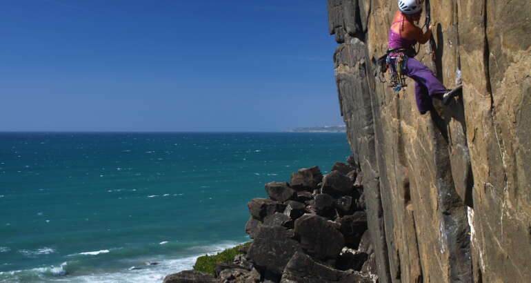 Inma Garrido aparece a la derecha de la foto escalando en una pared vertical. Al pie de vía se ven rocas y el mar que también ocupa toda la parte izquierda de la foto. Está tomada en Portugal en Casal de Pianos y el crédito de la foto es de Iván Jara