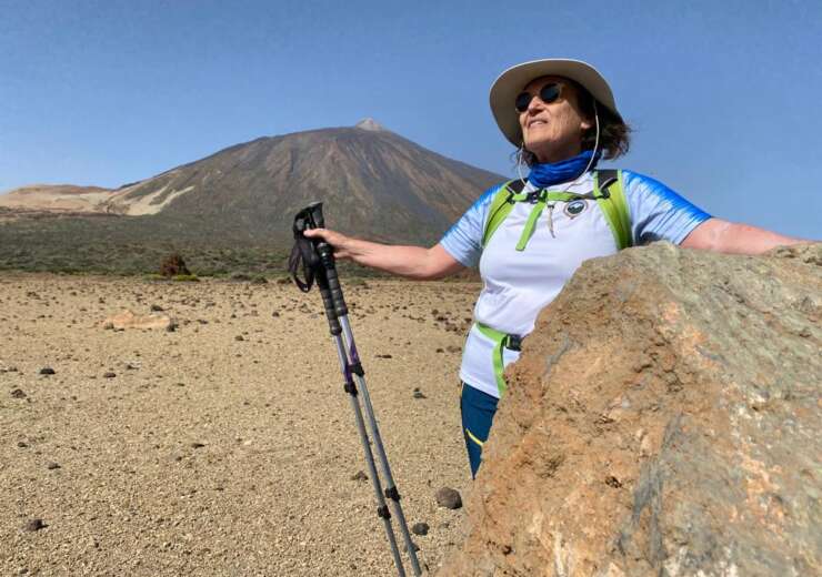 Josefina Suárez aparece en primer plano tras una roca en Parque Nacional del Teide. Su brazo izquierdo apoyado sobre la roca y el derecho con dos bastones hacia el suelo. Ella equipada con ropa de montaña, sombrero y gafas y mirando sonriente hacia arriba y al frente. Tras ella un gran llano de arena blanca y en el fondo el imponente Teide
