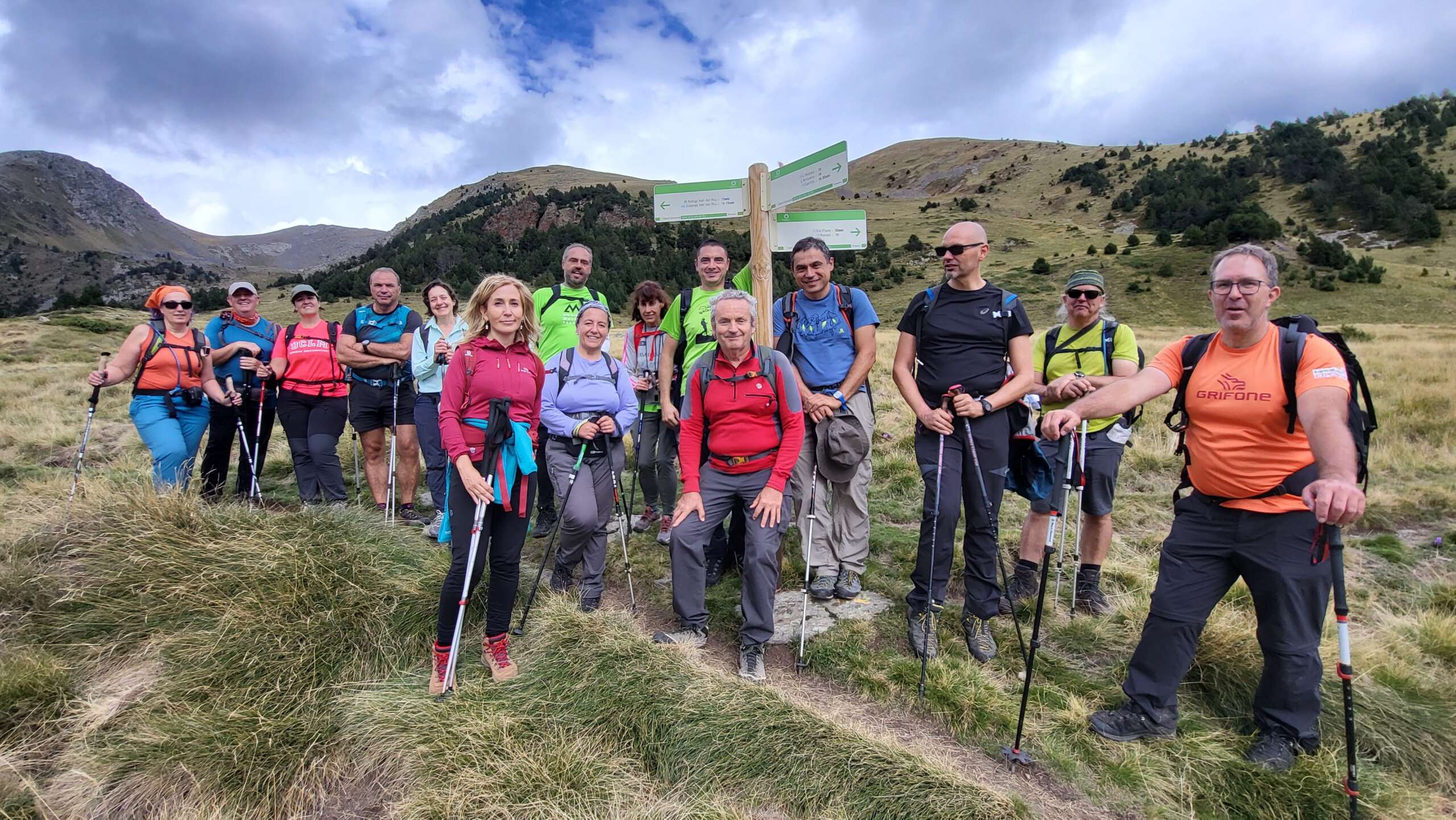 Fotografía en color que presenta a un grupo de senderistas parados en un poste direccional, ubicado en un paisaje de alta montaña, en una actividad del Andorra Walking Festival de 2022