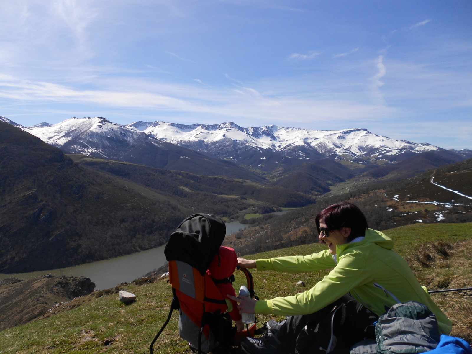 Patricia le sonríe a su hija, que está en una mochila roja de portabebés, lleva en la mano un biberón y en su rostro unas gafas de sol negras. Al fondo las montañas nevadas del valle de Nansa (Cantabria).