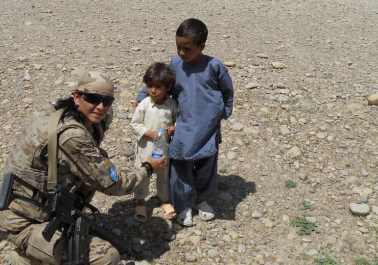 La Cabo 1ª Adelina Torres aparece a la izquierda de la foto con uniforme de campaña, agachada ofreciendo una botella de agua a dos niños mientras patrulla el aeropuerto de Qala-e-Naw en Afganistán.