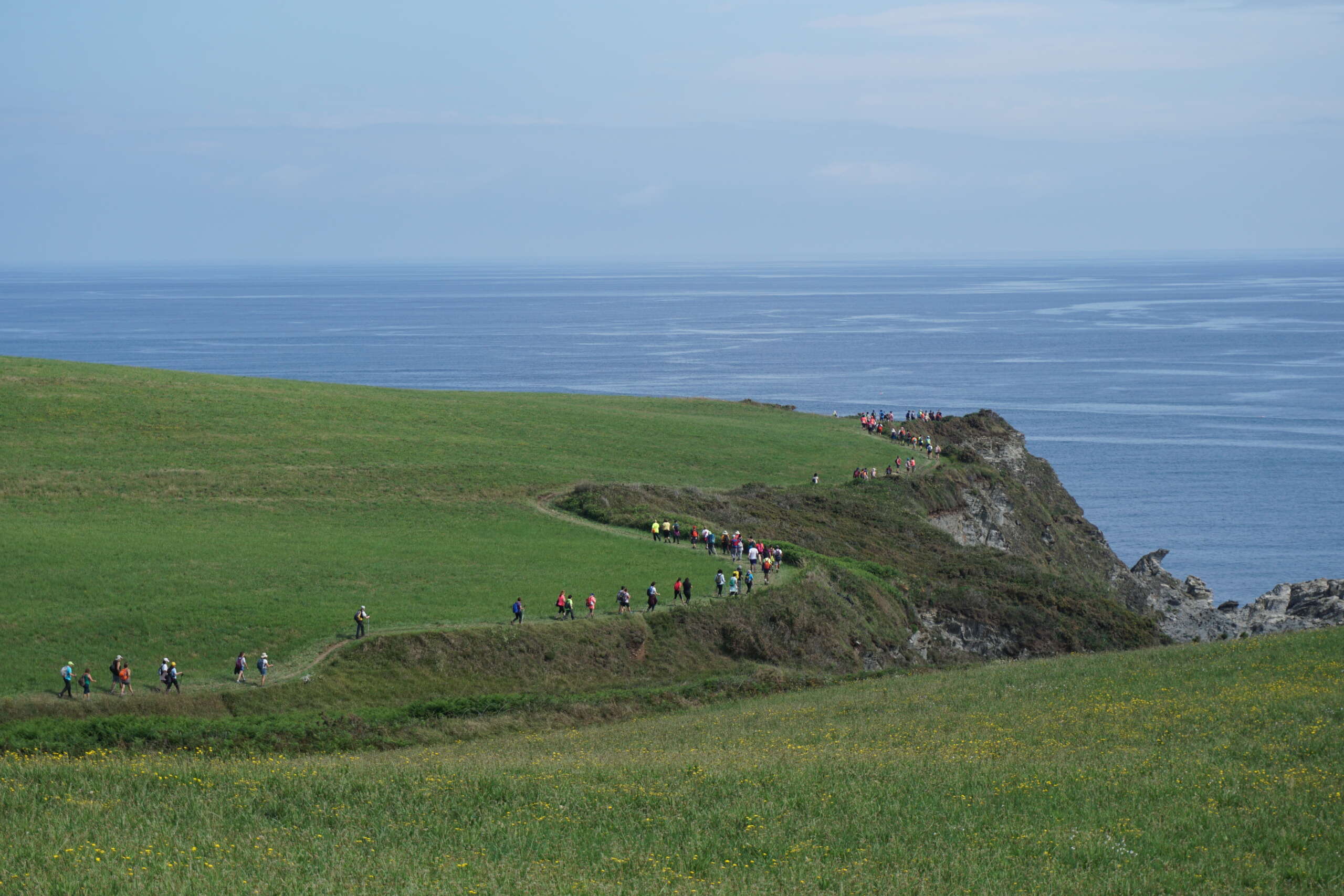 Foto en color que presenta a una serie de personas caminando por un camino entre los campos de hierbe y los acantilados sobre el mar