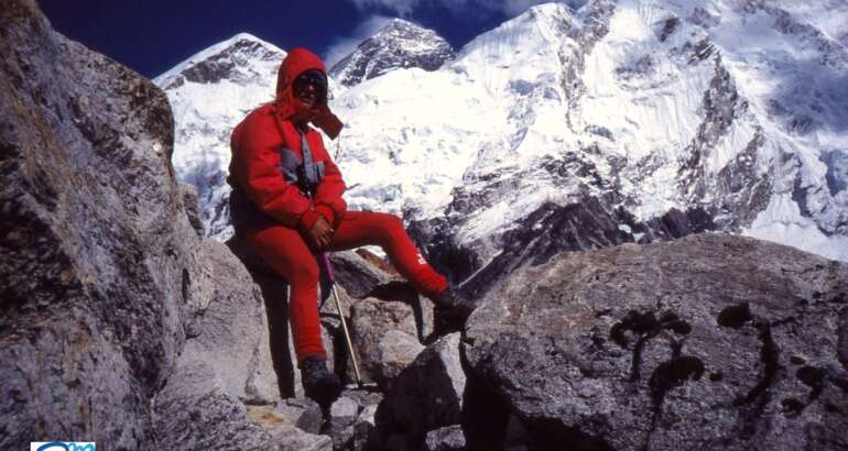 Reyes aparece sentada en unas grandes rocas con ropa de alta montaña roja, cubierta cabeza con capucha y gafas de nieve. Tras ella tres de las grandes montañas del Himalaya: Everest, Nuptse y Lhotse