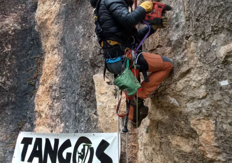 Un escalador en roca equipando una via. Con la imagen de una pancarta publicitaria de Tanogs en la Roca