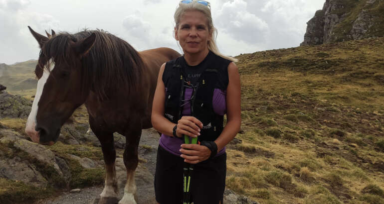 Peli aparece en medio de la foto en un paisaje montañoso con ropa de montaña y bastones de marcha nórdica. A su izquierda y justo detrás tiene un caballo que parece estar posando junto a ella.