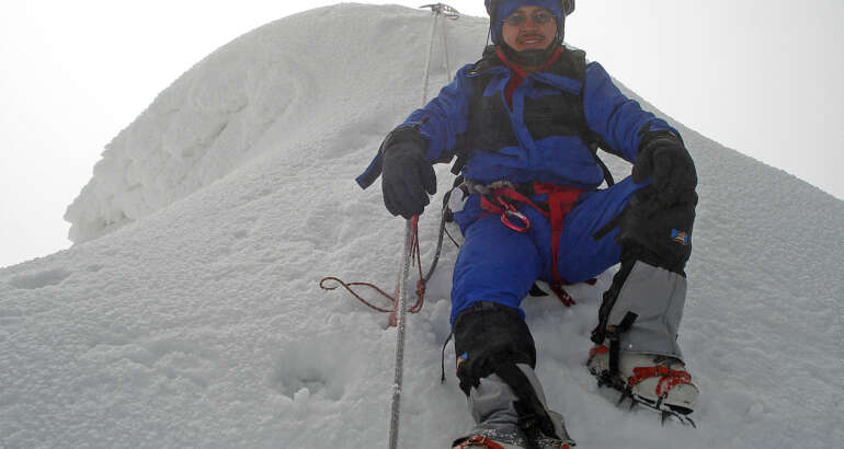 Christian Rodríguez en primer plano, sonriendo a la cámara, sentado en un montículo de hielo, mientras se asegura con una cuerda y varios piolets. Volcán Chimborazo de 6300 mts. Ecuador.