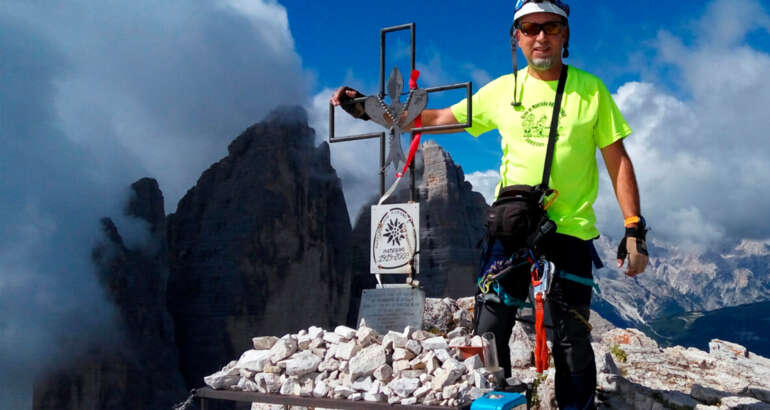 En la foto estoy tocando una cruz de forja con el símbolo de la flor de lis en el centro, en la cima de una montaña de los Dolomitas. Al fondo se ven las famosas Tres Cimas de Lavaredo