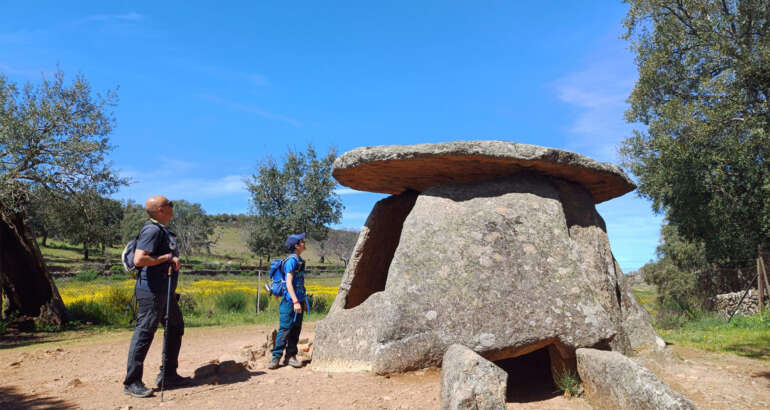 Ilustración a color que presenta a dos senderistas ante el dolmen de los Mellizos (Valencia de Alcántara-Cáceres), ubicado en una ruta de senderismo del Circuito Camina Extremadura, Liga Ibérica de Senderismo.